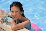 Woman leaning on edge of swimming pool, holding a drink, smiling at camera