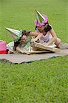 Two girls wearing party hats sitting on picnic blanket, opening a gift