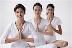 three women of mixed race sitting with legs crossed, arms in namaste, smiling at camera