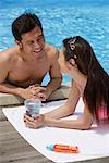 Couple looking at each other, man at the edge of swimming pool, woman lying down, holding glass of water