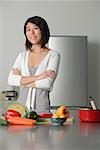 Jeune femme dans la cuisine, les bras croisés, les légumes sur la table en face d'elle