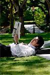 A man lies down and reads the newspaper in the park