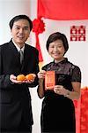 A couple look at the camera as they hold two oranges and a red envelope
