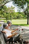 Couple in park, man reading newspaper, woman standing behind him looking over shoulder