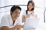 Couple in bedroom, man using laptop, looking at camera