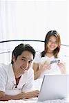 Couple in bedroom, man using laptop, smiling at camera