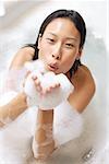 Woman in bathtub, blowing soap suds at camera