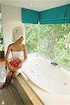 Woman in towel, sitting at edge of bath tub, holding bowl of flower petals