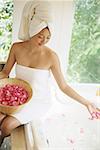 Woman in towel, sitting at edge of bath tub, throwing flower petals into water