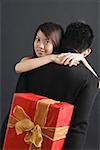Man holding gift behind his back, woman embracing him, looking over shoulder at camera