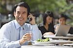 Homme d'affaires ayant un déjeuner au café en plein air, à l'aide de téléphone portable, souriant