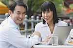 Businesswoman and businessman at outdoor cafe, with laptop, smiling at camera