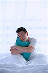 Man sitting in bed, embracing pillow, smiling at camera