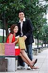 Couple looking at camera, man standing, woman sitting, shopping bags around them