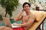 Man sitting by swimming pool, using laptop and mobile phone