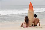 Couple sitting on beach, surfboard between them, looking out to sea