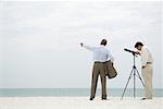 Two businessmen standing at the beach, one looking through telescope, the other pointing
