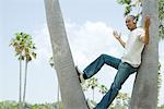 Man leaning against tree trunk listening to headphones, arms out, hands raised