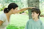 Boy leaning against tree trunk, his mother placing her hand on his head, both smiling