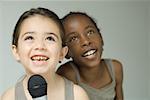 Two young girls singing into microphone together, both smiling and looking up