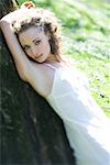 Young woman wearing slip, leaning against tree trunk, arm raised, smiling at camera