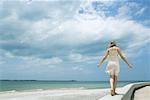 Young woman in sundress walking along low wall at the beach, arms out, rear view