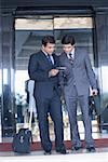 Two businessmen standing in front of the door of a hotel and looking at a mobile phone