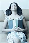 Young woman sitting on a couch with her eyes closed and holding a book