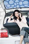 Young woman sitting in a car trunk and talking on a mobile phone
