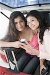Portrait of two young women sitting in a car trunk and holding a mobile phone