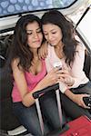 Two young women sitting in a car trunk and looking at a mobile phone