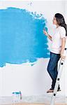 Side profile of a young woman painting a wall with a paint roller and leaning on a step ladder