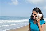 Jeune femme tenant une conque à son oreille et souriant sur la plage