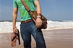 Mid section view of a young man carrying a shoulder bag and holding a pair of flip-flops on the beach