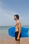 Side profile of a young man standing on the beach with a surfboard