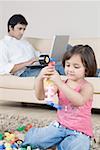 Girl playing with toys and her father working on a laptop in the background