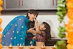 Side profile of a girl feeding a piece of burfi to her mother in a domestic kitchen