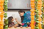 Young woman feeding a piece of burfi to her daughter in a domestic kitchen and smiling