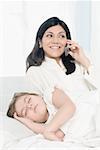 Mid adult woman talking on a mobile phone with her daughter sleeping beside her on the bed