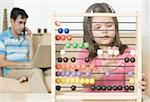 Girl playing with an abacus with her father talking on a mobile phone in the background