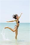 Young woman at the beach jumping, one leg up, arms raised