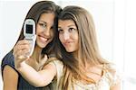 Teenage twin sisters photographing selves with cell phone, one smiling at camera