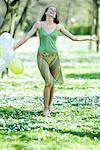 Young woman walking in meadow, holding balloons, arms out, smiling