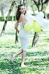 Young woman walking barefoot in meadow, holding balloons, smiling over shoulder at camera