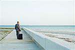 Businessman walking on sidewalk at the beach, pulling suitcase behind him, looking at view