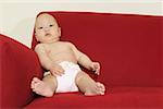 A baby seated on a red sofa and wearing a diaper.