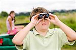 Girl sitting on car as brother uses binoculars