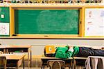 Boy lying on classroom desks with book over face