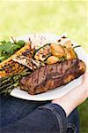 Femme tenant un plat de bœuf, légumes grillés & maïs en épi