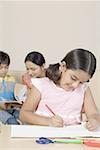 Girl coloring with crayon and her mother and brother sitting on a couch behind her